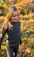 Child in a dark merino wool  long-sleeve shirt and pants standing in a field with trees in the background.