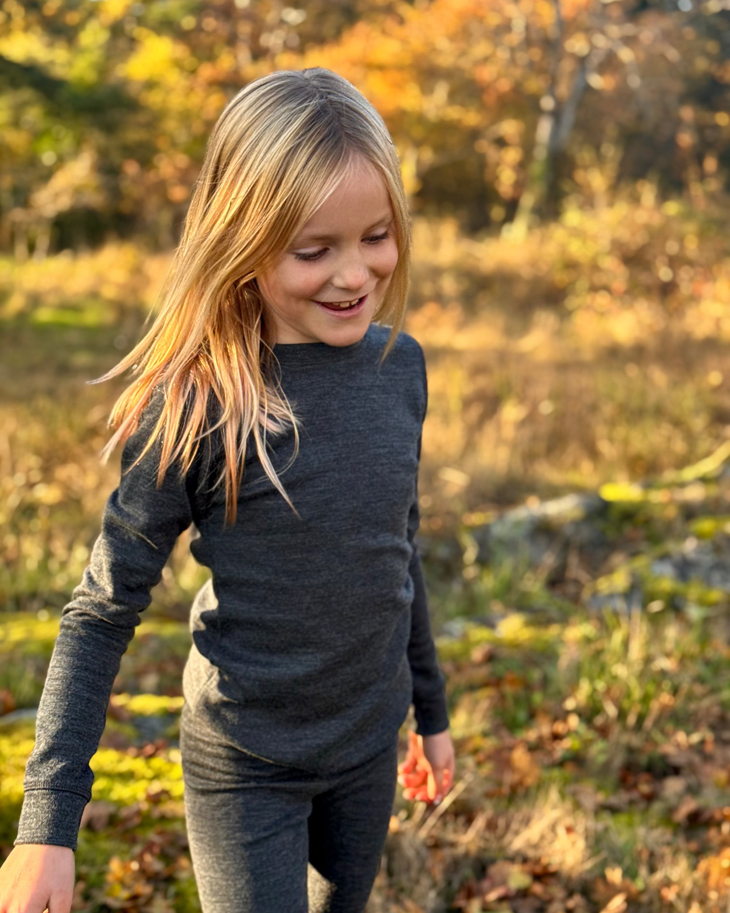 Child in a dark merino wool  long-sleeve shirt and pants standing in a field with trees in the background.