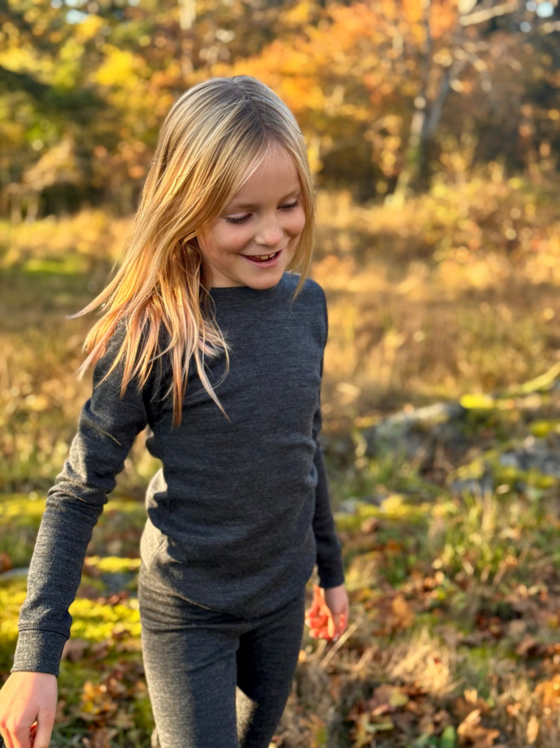 Child in a dark merino wool  long-sleeve shirt and pants standing in a field with trees in the background.