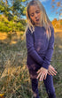 Child in merino wool purple top and bottoms standing in a field with trees and blue sky in the background