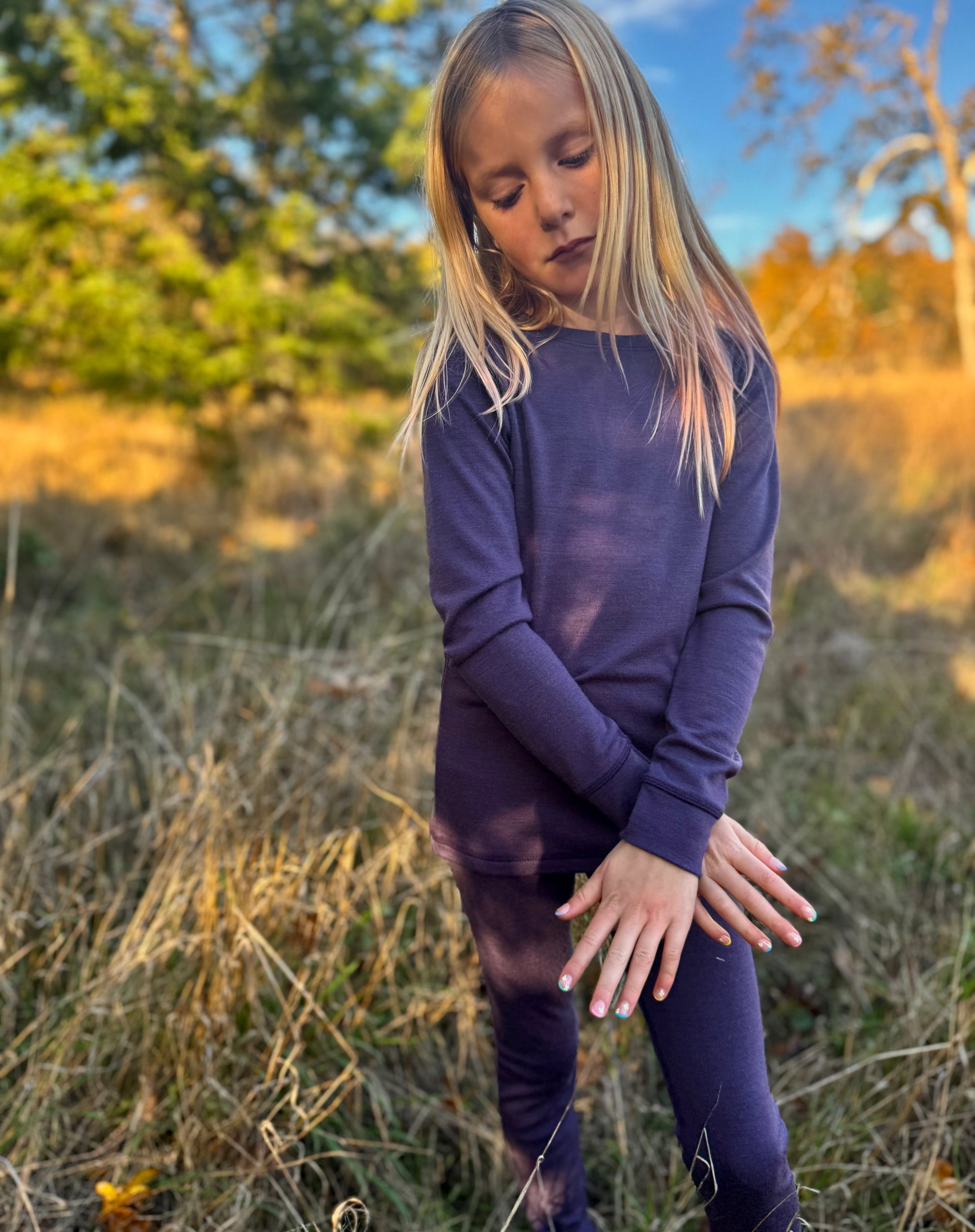 Child in merino wool purple top and bottoms standing in a field with trees and blue sky in the background