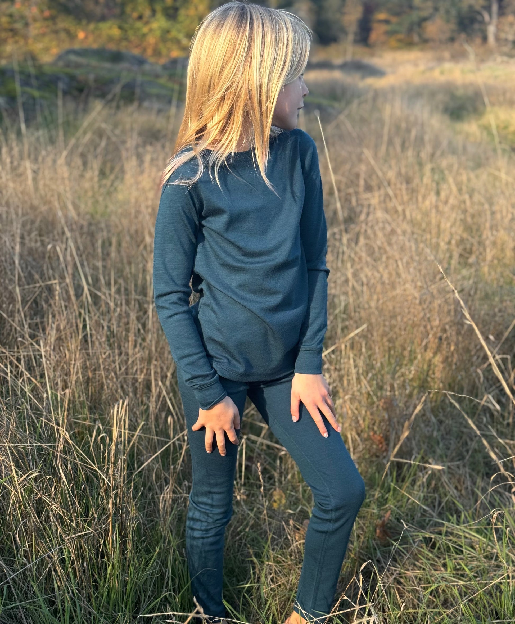 Child wearing a teal long-sleeve shirt Merino Wool Shirt and pants standing in a field with sunset lighting.